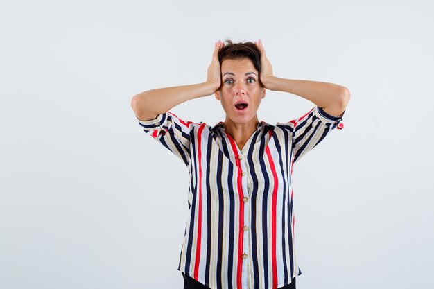 Mature woman clasping head with hands, keeping mouth open in striped shirt and looking surprised , front view.