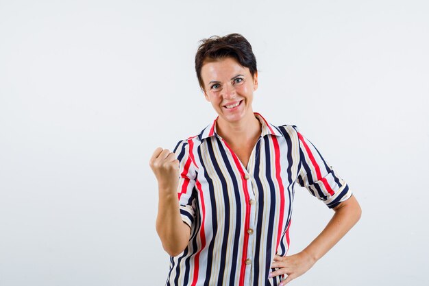 Mature woman clenching fist, holding hand on waist in striped blouse and looking confident. front view.