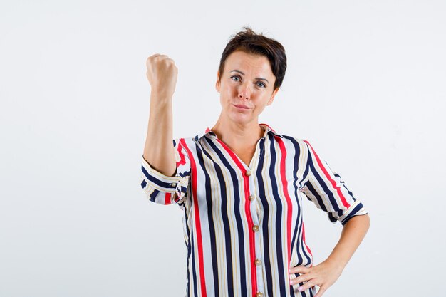 Зрелая женщина сжимает кулак, держит руку на талии в полосатой рубашке и выглядит уверенно, вид спереди.