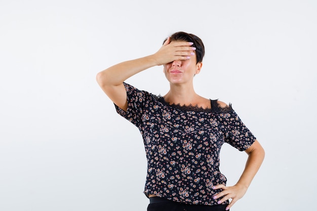 Mature woman covering eyes with hand, holding hand on waist in floral blouse, black skirt and looking cheery , front view.