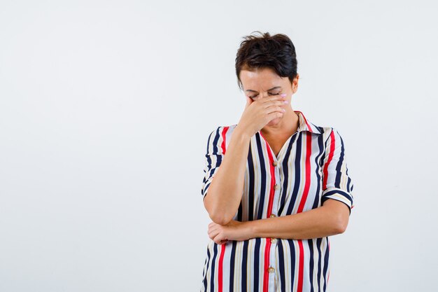 Mature woman covering mouth with hand, holding one hand under elbow in striped blouse and looking pensive. front view.