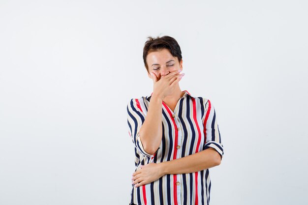 Mature woman covering mouth with hand, laughing, standing eyes closed in striped shirt and looking cheery. front view.