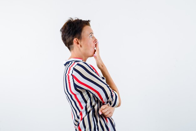 Mature woman covering mouth with hand in striped blouse and looking shocked , front view.