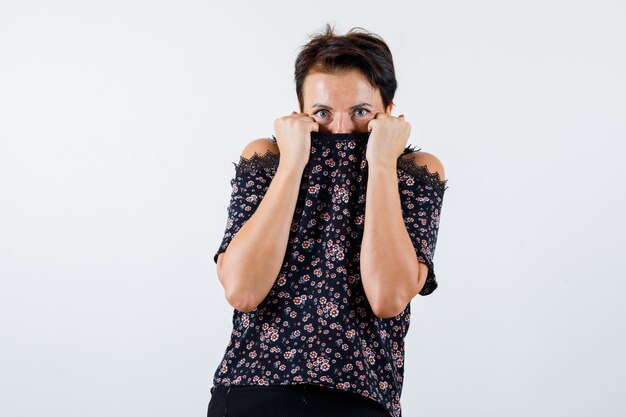 Mature woman in floral blouse, black skirt hiding behind collar and looking scared , front view.