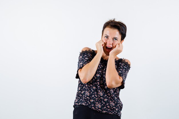 Mature woman in floral blouse, black skirt holding hands on cheeks, keeping mouth open and looking cheery , front view.