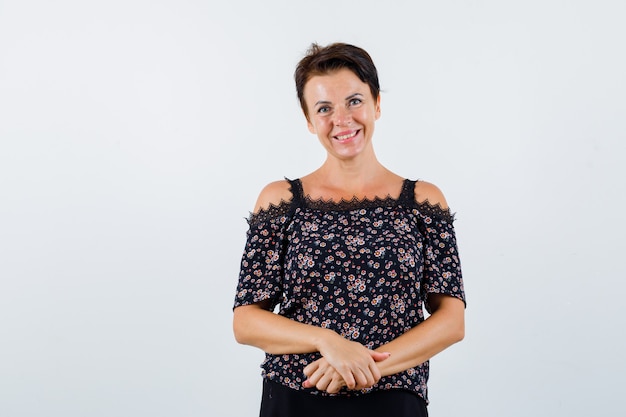 Mature woman in floral blouse, black skirt holding hands in front of her and looking jolly , front view.