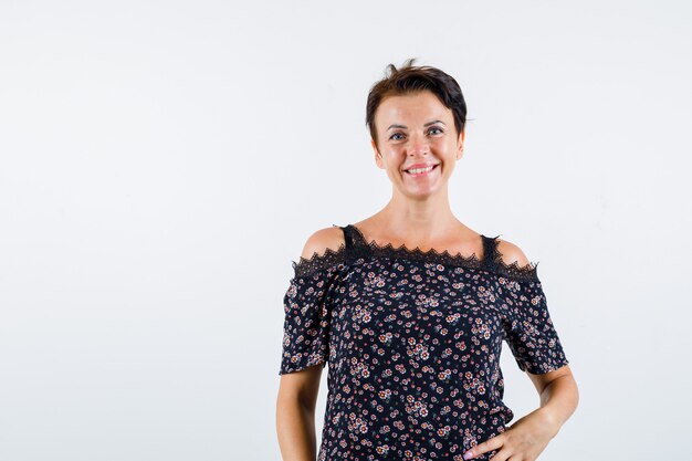 Free photo mature woman in floral blouse, black skirt holding one hand on waist, smiling and looking jolly , front view.