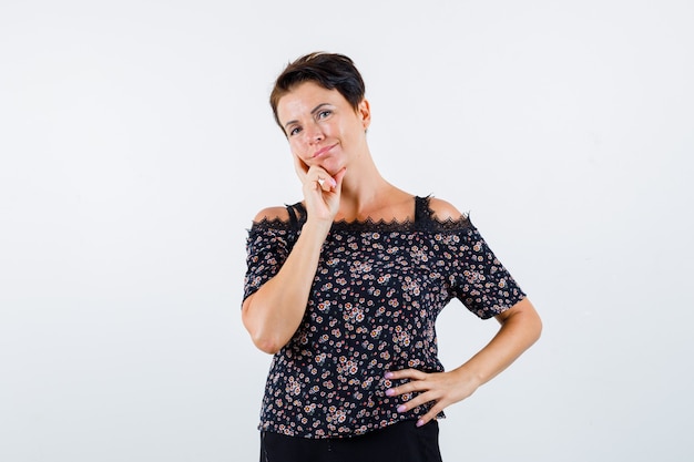 Mature woman in floral blouse, black skirt leaning cheek on index finger, thinking about something and looking pensive , front view.