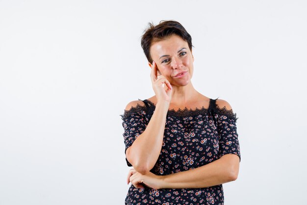 Mature woman in floral blouse, black skirt leaning cheek on palm, curving lips, thinking about something and looking pensive , front view.