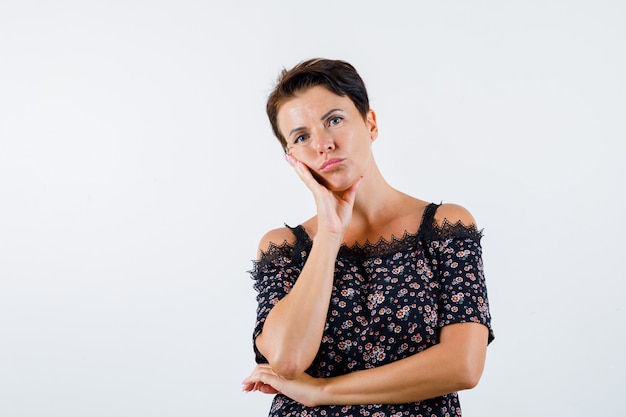 Mature woman in floral blouse, black skirt leaning cheek on palm, thinking about something and looking pensive , front view.