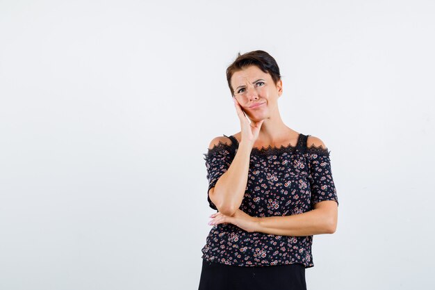 Mature woman in floral blouse and black skirt leaning cheek on palm, thinking about something and looking pensive , front view.