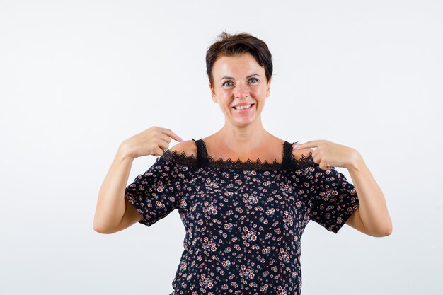 Mature woman in floral blouse, black skirt pointing at herself with index fingers and looking cheery , front view.