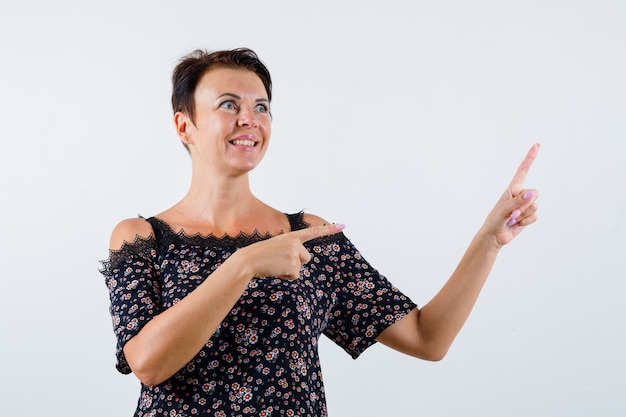 Mature woman in floral blouse, black skirt pointing right and up with index finger, looking upward and looking cheery , front view.