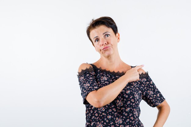 Mature woman in floral blouse, black skirt pointing behind with index finger, holding hand on waist and looking serious , front view.