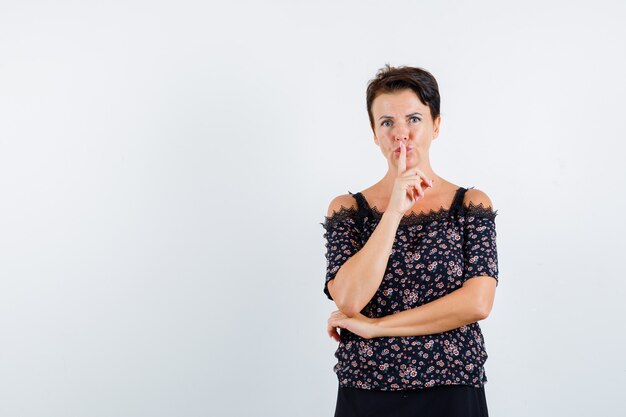 Mature woman in floral blouse and black skirt showing silence gesture and looking serious , front view.