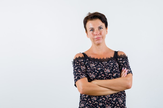 Mature woman in floral blouse, black skirt standing arms crossed and looking confident , front view.