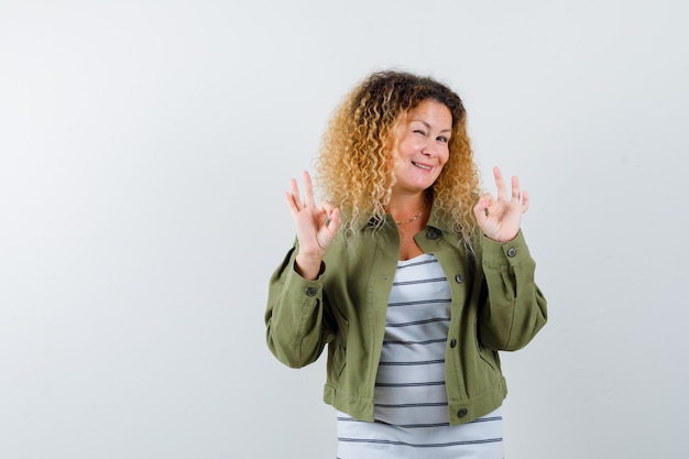 Mature woman in green jacket, t-shirt showing ok signs and looking pleased , front view.