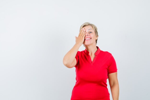 Mature woman holding hand on eye in red t-shirt and looking curious
