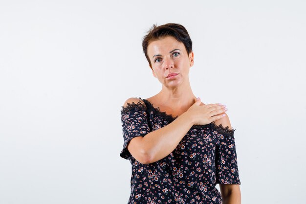 Mature woman holding hand on shoulder in floral blouse, black skirt and looking serious. front view.