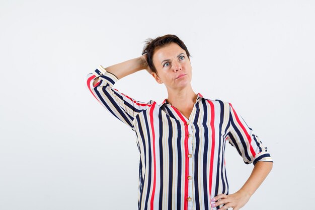 Зрелая женщина, держащая руку на талии, держащая одну руку на голове в полосатой блузке и задумчивая, вид спереди.