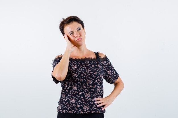 Mature woman holding hand on waist, leaning cheek on palm, looking away in floral blouse, black skirt and looking pensive. front view.