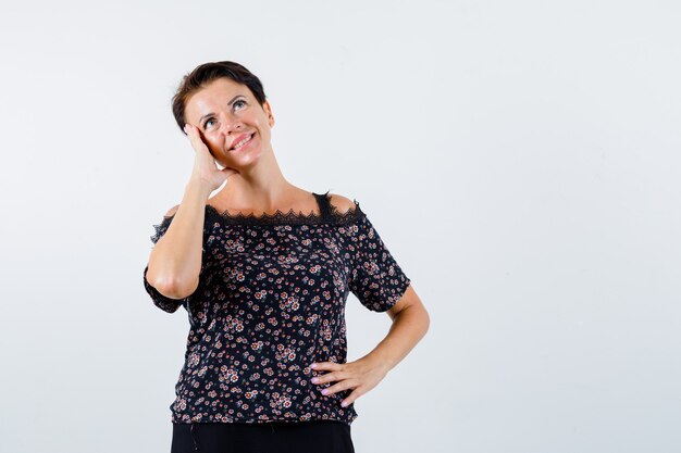 Mature woman holding hand on waist, leaning cheek on palm, smiling in floral blouse, black skirt and looking pensive. front view.