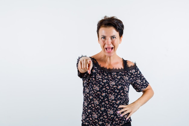 Mature woman holding hand on waist, pointing at camera, keeping mouth open in floral blouse, black skirt and looking jolly. front view.