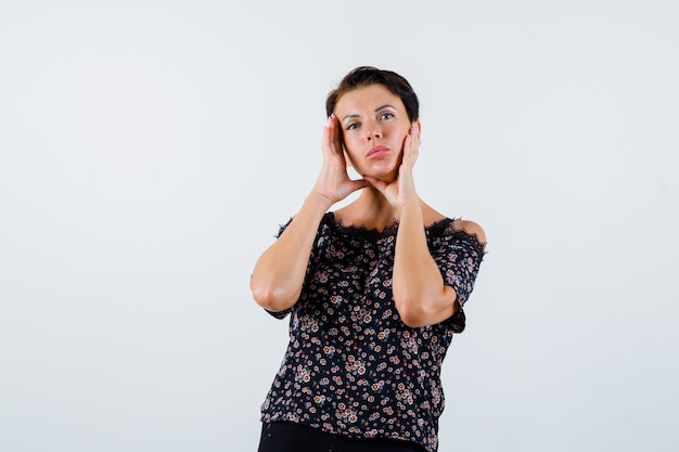 Mature woman holding hands on cheeks in floral blouse, black skirt and looking charming. front view.