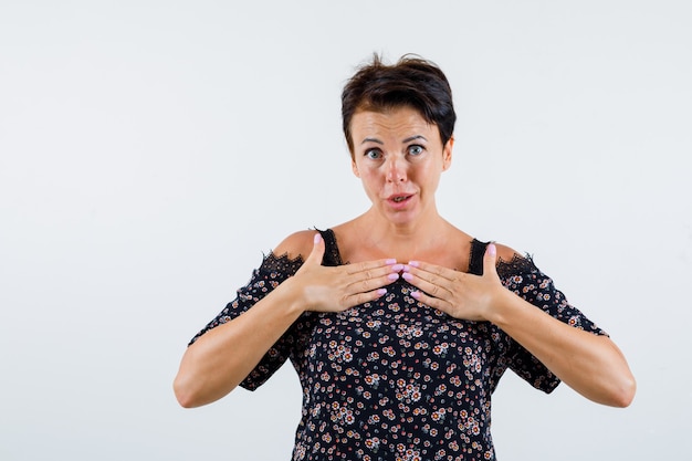 Mature woman holding hands over chest in floral blouse, black skirt and looking surprised. front view.