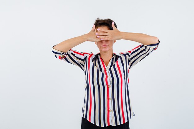 Mature woman holding hands on eyes in striped shirt and looking calm , front view.