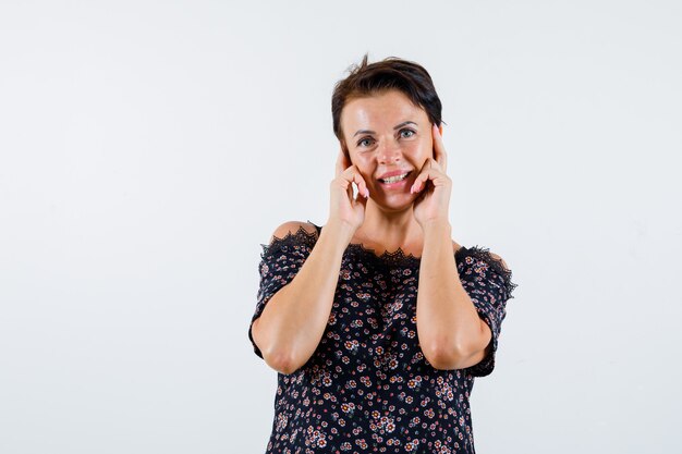 Mature woman holding index fingers on cheeks in floral blouse, black skirt and looking cheery. front view.