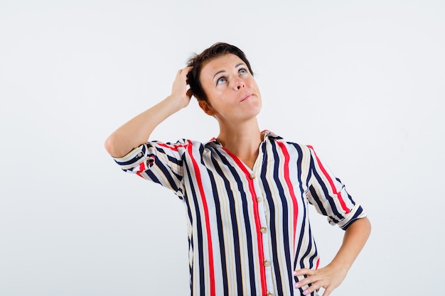 Mature woman holding one hand on head, another hand on waist, looking upward in striped shirt and looking pensive , front view.