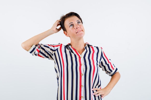 Mature woman holding one hand on head, another hand on waist in striped shirt and looking pensive. front view.