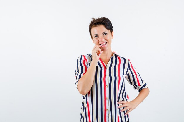 Mature woman holding one hand on waist, biting fingers in striped shirt and looking happy , front view.