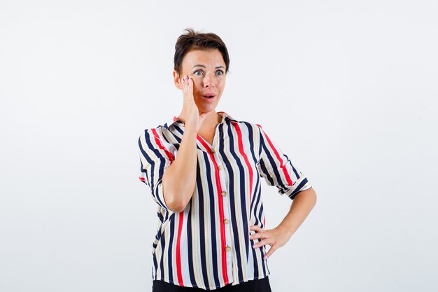 Mature woman holding one hand on waist, keeping another hand near mouth as calling in striped shirt and looking surprised. front view.