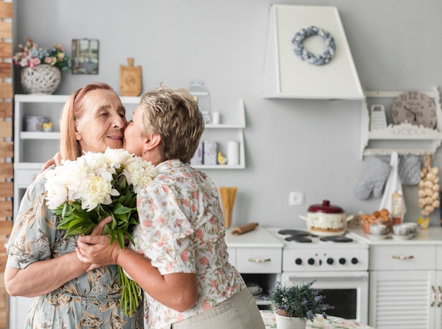 Mature woman kissing her mother holding white flower bouquet at home