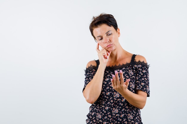 Mature woman leaning cheek on hand, looking at hand as holding something in it in floral blouse, black skirt and looking focused. front view.