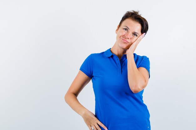 Зрелая женщина, опираясь щекой на ладонь в синей футболке и глядя задумчиво, вид спереди.