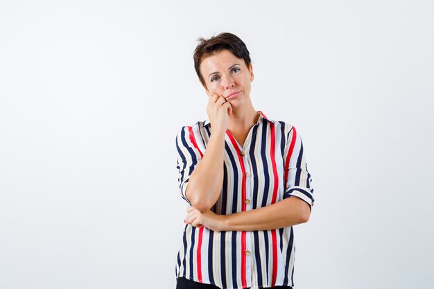 Mature woman leaning cheek on palm, holding one hand under elbow in striped shirt and looking pensive. front view.