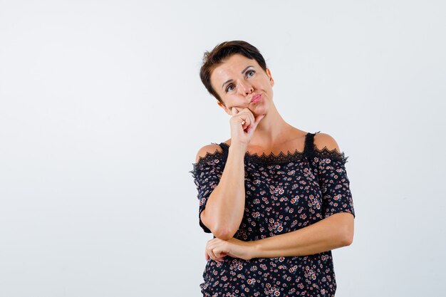 Mature woman leaning cheek on palm, thinking about something in floral blouse, black skirt and looking pensive , front view.