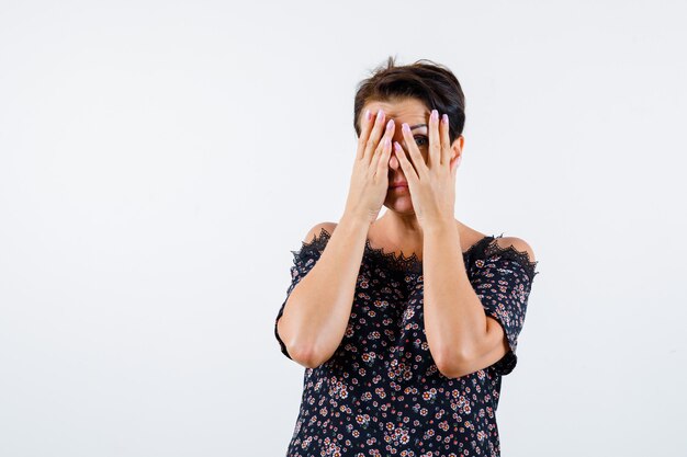 Mature woman looking through fingers in floral blouse, black skirt and looking serious , front view.