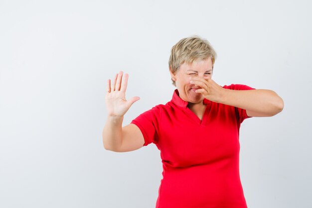 Mature woman pinching nose due to bad smell in red t-shirt and looking disgusted.