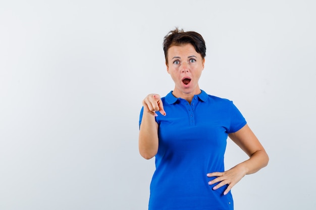 Mature woman pointing at camera in blue t-shirt and looking surprised. front view.