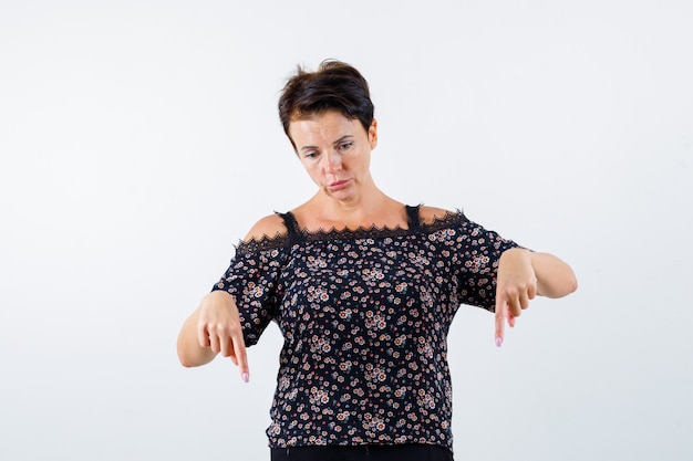 Mature woman pointing down with index fingers in floral blouse, black skirt and looking serious. front view