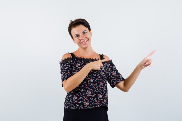 Mature woman pointing right with index fingers in floral blouse, black skirt and looking cheery , front view.