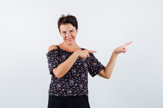 Mature woman pointing right with index fingers in floral blouse, black skirt and looking jolly , front view.