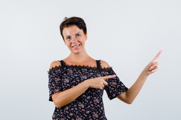 Зрелая женщина, указывающая вправо указательными пальцами в цветочной блузке, черной юбке и веселый вид спереди.