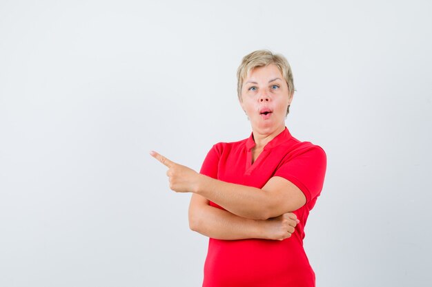 Mature woman pointing at upper left corner in red t-shirt and looking amazed.