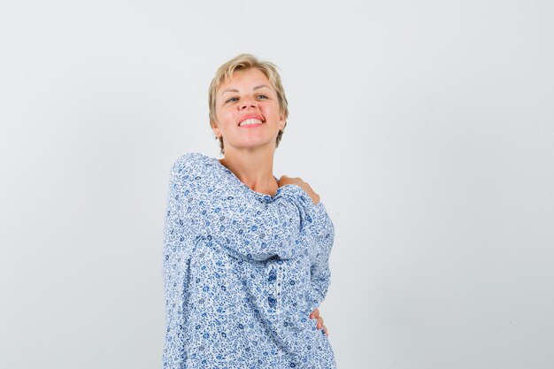 Mature woman posing while holding hand on shoulder in shirt and looking cheery.