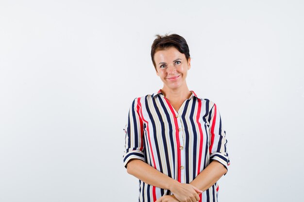 Mature woman posing with clasped hands, smiling in striped blouse and looking sanguine. front view.